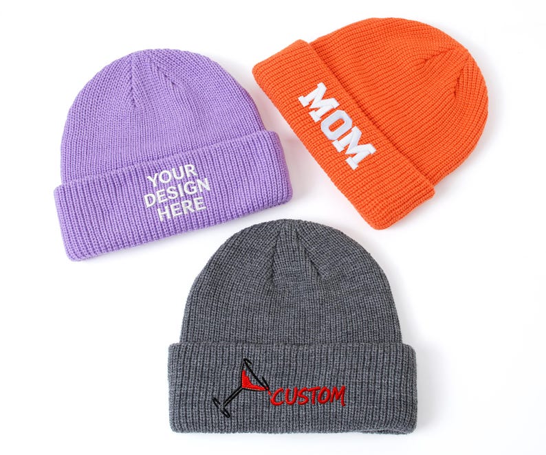 May include: Three knit beanies in lavender, orange, and dark gray. The lavender beanie features white text reading YOUR DESIGN HERE. The orange beanie has white text reading MOM. The dark gray beanie displays a red cocktail glass icon and the red text CUSTOM.