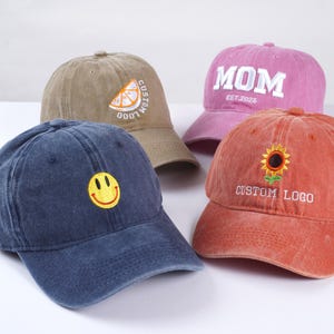 May include: Four different colored baseball caps with embroidered designs. The caps are brown, pink, blue, and orange. The designs include a lemon slice, the word "MOM" with the date "EST. 2025", a smiley face, and a sunflower with the text "CUSTOM LOGO".