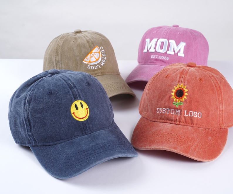 May include: Four different colored baseball caps with embroidered designs. The caps are brown, pink, blue, and orange. The designs include a lemon slice, the word "MOM" with the date "EST. 2025", a smiley face, and a sunflower with the text "CUSTOM LOGO".