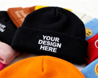 May include: A black beanie with the text "YOUR DESIGN HERE" in white. The beanie is surrounded by other beanies in various colors, including orange, brown, and pink. The beanies are made of a knitted material.