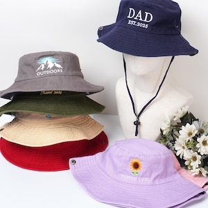 May include: A collection of bucket hats in various colors, including navy blue, gray, red, and lavender. The navy hat has "DAD EST.2025" embroidered on it. Other hats feature mountain and sunflower designs, and the text "OUTDOORS", "Travel 2025", and "CUSTOM LOGO".
