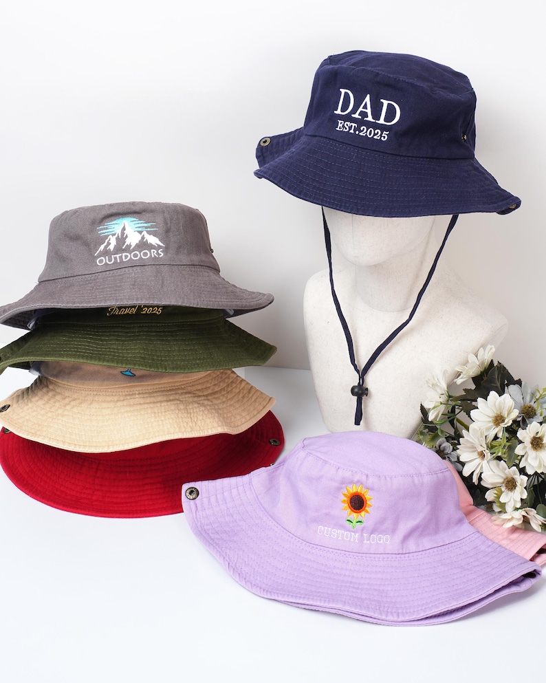 May include: A collection of bucket hats in various colors, including navy blue, gray, red, and lavender. The navy hat has "DAD EST.2025" embroidered on it. Other hats feature mountain and sunflower designs, and the text "OUTDOORS", "Travel 2025", and "CUSTOM LOGO".