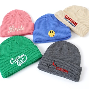 May include: Five knit beanies in pink, blue, cream, green, and charcoal gray. The pink beanie features the word Bride, the blue one has a yellow smiley face, the cream one says CUSTOM YOUR DESIGN, the green one says CUSTOM TEXT, and the gray one has a martini glass icon with the word CUSTOM.