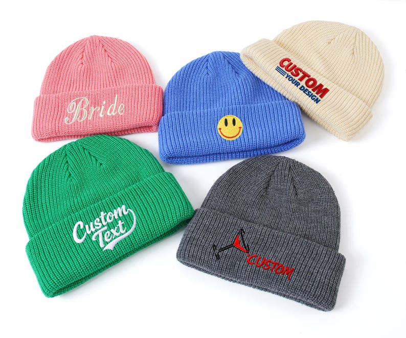 May include: Five knit beanies in pink, blue, cream, green, and charcoal gray. The pink beanie features the word Bride, the blue one has a yellow smiley face, the cream one says CUSTOM YOUR DESIGN, the green one says CUSTOM TEXT, and the gray one has a martini glass icon with the word CUSTOM.