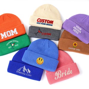 May include: A collection of eight knit beanies in orange, green, cream, dark gray, purple, brown, blue, and pink. Each beanie features embroidered text or graphics, including 'MOM', 'CUSTOM YOUR DESIGN', 'Bride', a smiley face, a mountain, and a sunflower.