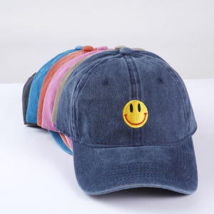 May include: A stack of six baseball caps in various colors, including blue, pink, and brown. The top cap is dark blue with a yellow smiley face embroidered on the front.