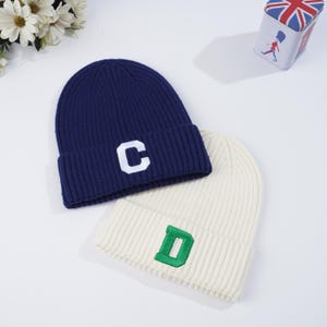 Może przedstawiać: Dwie dzianinowe czapki, jedna granatowa z białym "C" i druga kremowa z zielonym "D". Czapki są prążkowane i złożone na brzegu. Tło jest białe.