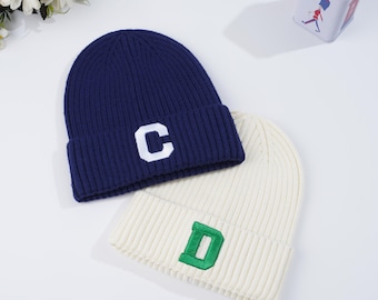 May include: Two knit beanies, one navy blue with a white "C" and the other cream-colored with a green "D". The beanies are ribbed and folded at the brim. The background is white.