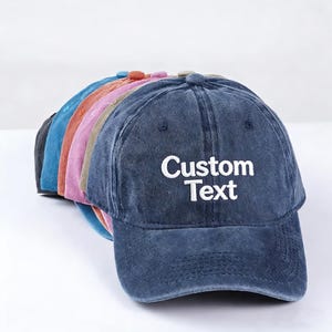 May include: A stack of six baseball caps in various colors including dark blue, light blue, orange, pink, tan, and black. The front-most dark blue cap features the white embroidered text Custom Text centered on the front panel above the brim.