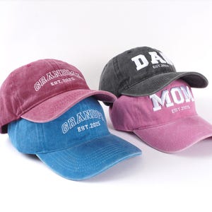 May include: Three baseball caps with different embroidered text. The first cap is red with "GRANDPA EST. 2025" in white. The second cap is blue with "GRANDPA EST. 2025" in white. The third cap is pink with "MOM EST. 2025" in white. The cap on top is black with "DAD EST. 2025" in white.