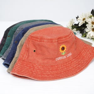 Könnte beinhalten: Ein Stapel aus sechs verschiedenen Canvas-Bucket-Hüten in verschiedenen Farben. Der oberste Hut ist orange mit einer Sonnenblume und dem gestickten Text "CUSTOM LOGO" auf der Vorderseite.