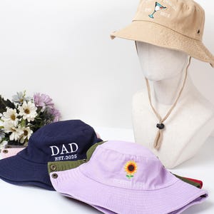 May include: A collection of bucket hats in various colors. One tan hat features a cocktail design and the text "CUSTOM DESIGN EST. 2025." A navy blue hat displays "DAD EST.2025." A lavender hat has a sunflower and "CUSTOM LOGO" embroidered on it.