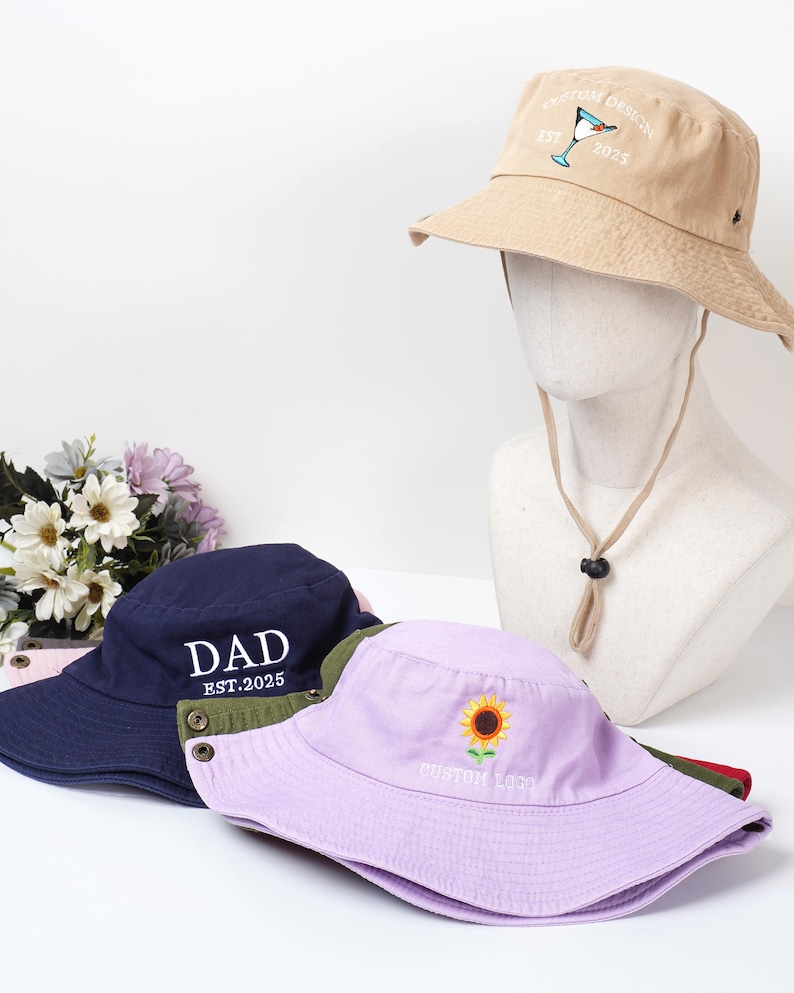 May include: A collection of bucket hats in various colors. One tan hat features a cocktail design and the text "CUSTOM DESIGN EST. 2025." A navy blue hat displays "DAD EST.2025." A lavender hat has a sunflower and "CUSTOM LOGO" embroidered on it.