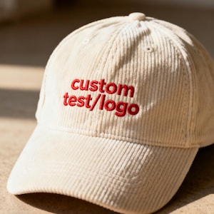 Peut inclure: Casquette de baseball en velours côtelé crème avec le texte "custom test/logo" brodé en rouge. La casquette a une visière incurvée et une couronne structurée. Le chapeau est en tissu doux et texturé.