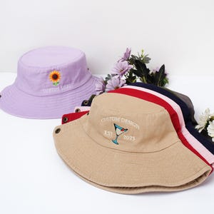 May include: Several bucket hats in various colors are displayed. A light purple hat features a sunflower embroidery and the text "CUSTOM LOGO". A tan hat has a cocktail glass design and the text "CUSTOM DESIGN EST 2025". Other hats are visible with red, pink, navy, and gray stripes.