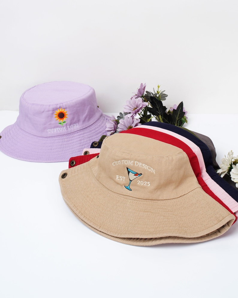 May include: Several bucket hats in various colors are displayed. A light purple hat features a sunflower embroidery and the text "CUSTOM LOGO". A tan hat has a cocktail glass design and the text "CUSTOM DESIGN EST 2025". Other hats are visible with red, pink, navy, and gray stripes.