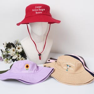 May include: Several bucket hats in different colours are displayed. A red hat with the text "your text/logo here" is on a mannequin head. Other hats include a lilac hat with a sunflower and a beige hat with a cocktail design.