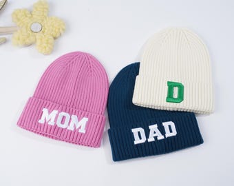 May include: Three knitted beanies in pink, white, and blue with the words "MOM", "DAD", and the letter "D" embroidered on them.