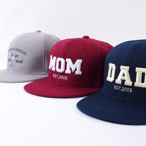 Puede incluir: Tres gorras de béisbol. La primera es gris con un diseño de cóctel y el texto "CUSTOM DESIGN EST. 2025". La segunda es granate con "MOM EST. 2025". La tercera es azul marino con "DAD EST. 2025".
