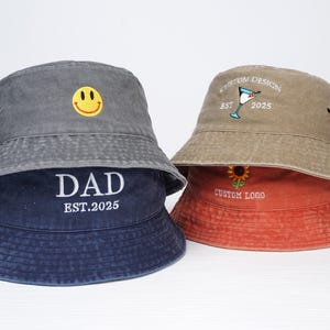 May include: Three different styles of bucket hats. The first hat is gray with a yellow smiley face embroidered on the front. The second hat is beige with a martini glass and the text "Custom Design Est. 2025" embroidered on the front. The third hat is red with a sunflower and the text "Custom Logo" embroidered on the front.
