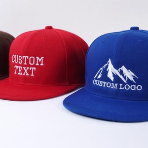 Puede incluir: Tres gorras de béisbol con viseras planas. La primera es marrón, la segunda es roja con "CUSTOM TEXT" bordado en blanco, y la tercera es azul con un gráfico de montaña y "CUSTOM LOGO" bordado en blanco.