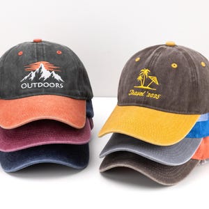 May include: A stack of six baseball caps in various colours. Two caps have the text "OUTDOORS" with a mountain graphic. The other four caps have the text "Travel '2025" with a palm tree and umbrella graphic. The caps have a distressed appearance.