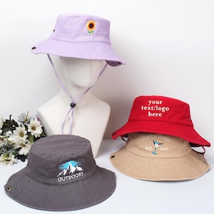 May include: Four bucket hats in various colors: lavender with a sunflower, red with text, gray with a mountain design, and tan with a sailboat. Each hat has a brim and adjustable chin strap.