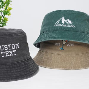 Könnte beinhalten: Zwei Bucket-Hats, einer schwarz mit dem Text "CUSTOM TEXT" vorne bestickt, und einer grün mit dem Text "CUSTOM LOGO" vorne und "EST 2025" hinten bestickt.