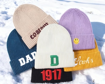 May include: A collection of six ribbed knit beanies in various colors including beige with the text 'costom', dark blue with 'DAD', white with a green 'D', black with red '1917', yellow with 'Stroh's', and purple with a yellow smiley face patch.