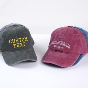 May include: Two adjustable baseball caps. One is dark gray with yellow embroidered text that says "CUSTOM TEXT". The other is burgundy with white embroidered text that says "GRANDMA EST. 2025".