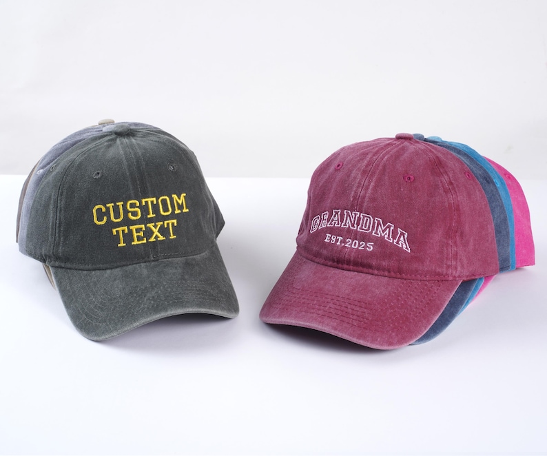 May include: Two adjustable baseball caps. One is dark gray with yellow embroidered text that says "CUSTOM TEXT". The other is burgundy with white embroidered text that says "GRANDMA EST. 2025".