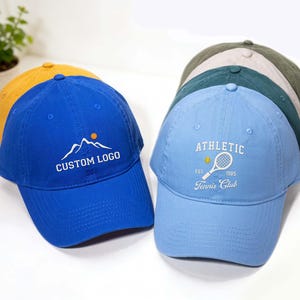 Puede incluir: Una colección de gorras de béisbol en varios colores, incluyendo azul, amarillo y verde. Una gorra azul presenta un logotipo de montaña y el texto "CUSTOM LOGO". Otra gorra azul claro muestra "ATHLETIC Tennis Club" con un gráfico de raqueta de tenis.