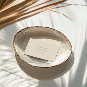 Mockup of a business card placed in a ceramic bowl with a rustic, speckled glaze sitting on a white surface. Sunlight casts soft shadows of palm leaves across the scene, creating a warm, natural aesthetic.
