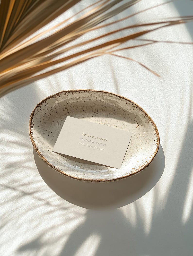 Mockup of a business card placed in a ceramic bowl with a rustic, speckled glaze sitting on a white surface. Sunlight casts soft shadows of palm leaves across the scene, creating a warm, natural aesthetic.