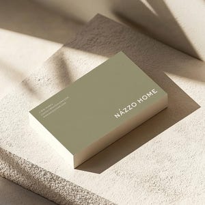 Op de afbeelding: Een groene visitekaartje met de tekst "N&Aacute;ZZO HOME" in witte letters. De kaart ligt op een getextureerd, lichtbeige oppervlak.
