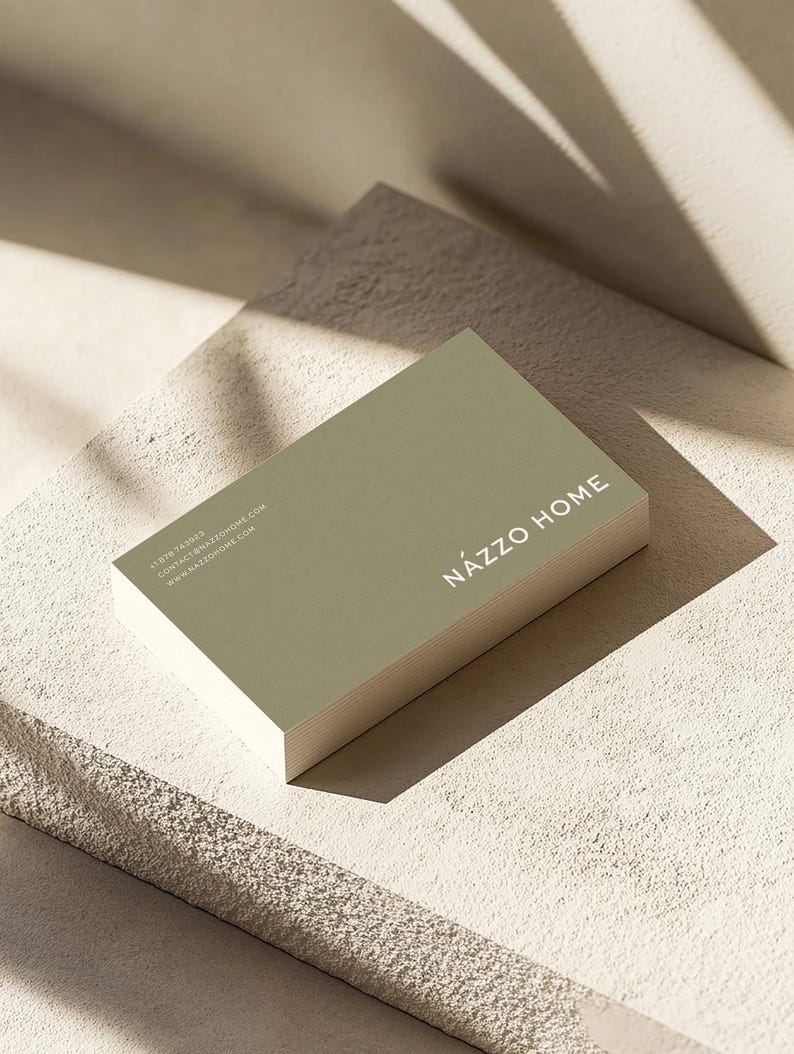 Op de afbeelding: Een groene visitekaartje met de tekst "N&Aacute;ZZO HOME" in witte letters. De kaart ligt op een getextureerd, lichtbeige oppervlak.
