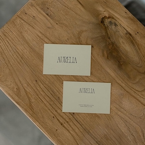 May include: Two business cards with the name "AURELIA" printed in black on a light-colored background. One card includes contact information. The cards are on a wooden surface.