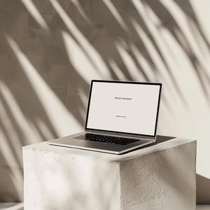 Peut inclure: Un ordinateur portable argenté avec un écran blanc vide affichant le texte "DEVICE MOCKUP" sur un piédestal en béton blanc. L'arrière-plan est un mur beige clair avec des ombres d'une feuille de palmier.