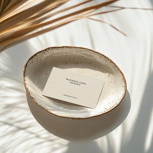 Mockup of a business card placed in a ceramic bowl with a rustic, speckled glaze sitting on a white surface. Sunlight casts soft shadows of palm leaves across the scene, creating a warm, natural aesthetic.