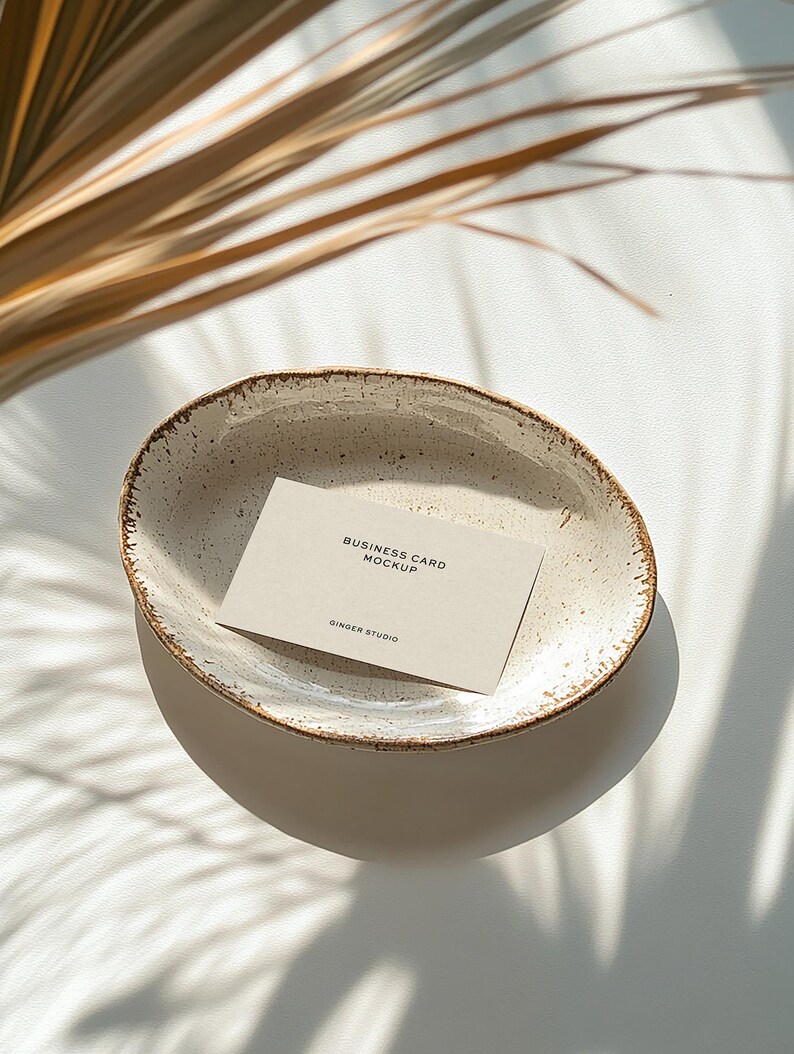 Mockup of a business card placed in a ceramic bowl with a rustic, speckled glaze sitting on a white surface. Sunlight casts soft shadows of palm leaves across the scene, creating a warm, natural aesthetic.