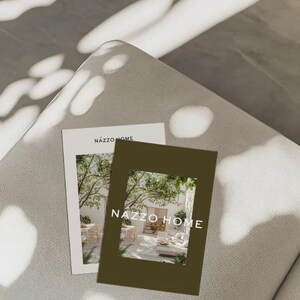 May include: Two brochures with the text "NAZZO HOME" are displayed on a light gray surface. One brochure is white with a photo of an outdoor living space. The other is a dark olive green with a photo of an outdoor living space.