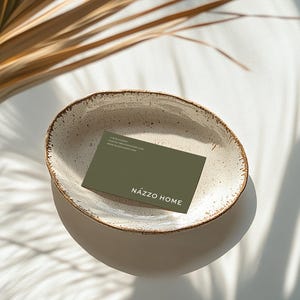 Mockup of a business card placed in a ceramic bowl with a rustic, speckled glaze sitting on a white surface. Sunlight casts soft shadows of palm leaves across the scene, creating a warm, natural aesthetic.