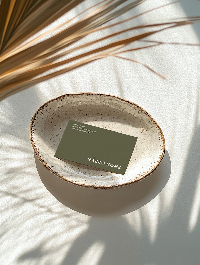 Mockup of a business card placed in a ceramic bowl with a rustic, speckled glaze sitting on a white surface. Sunlight casts soft shadows of palm leaves across the scene, creating a warm, natural aesthetic.