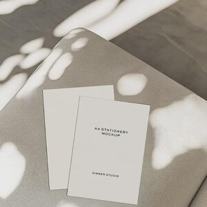 May include: Two white A4 stationery mockups on a gray surface. One mockup is blank, and the other has the text "A4 STATIONERY MOCKUP" and "GINGER STUDIO" printed on it. The scene is lit with natural light and shadows.