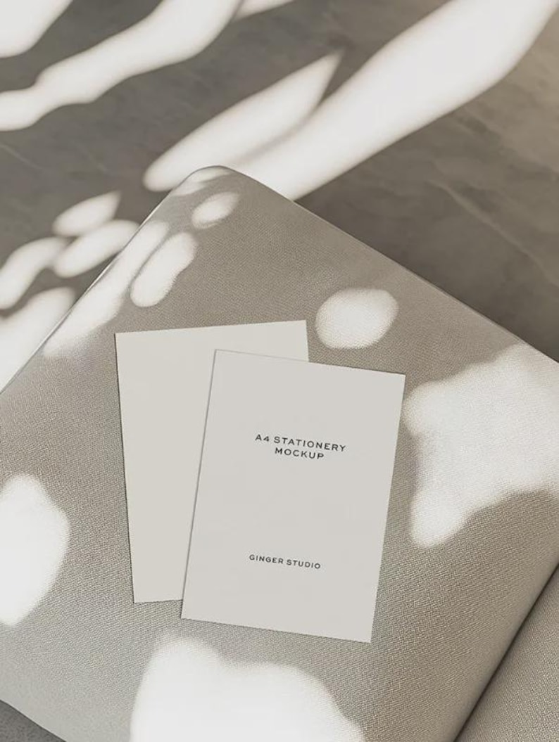 May include: Two white A4 stationery mockups on a gray surface. One mockup is blank, and the other has the text "A4 STATIONERY MOCKUP" and "GINGER STUDIO" printed on it. The scene is lit with natural light and shadows.