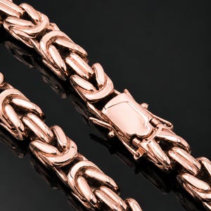 Könnte beinhalten: Eine byzantinische Kettenhalskette aus Roségold mit einer Reihe ineinandergreifender, gewebter Metallglieder, die ein strukturiertes, zylindrisches Aussehen erzeugen. Die Kette verfügt über einen sicheren, polierten rechteckigen Kastenverschluss.