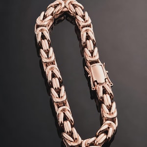 Könnte beinhalten: Ein Armband mit byzantinischer Kette in Roségold-Optik, bestehend aus einer Reihe ineinandergreifender, abgerundeter Metallglieder, die eine texturierte, gewebte Optik erzeugen. Das Armband wird mit einem rechteckigen Kastenverschluss geschlossen.