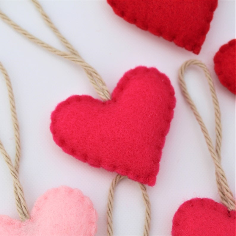 May include: A set of felt heart ornaments in pink and red, each with a twine loop for hanging. The hearts are handmade and have a rustic, charming look.