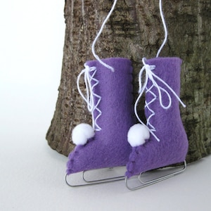 Pode incluir: Dois enfeites de patins de gelo de feltro roxo com pompons brancos e cadarços brancos. Os enfeites estão pendurados em um galho de árvore.