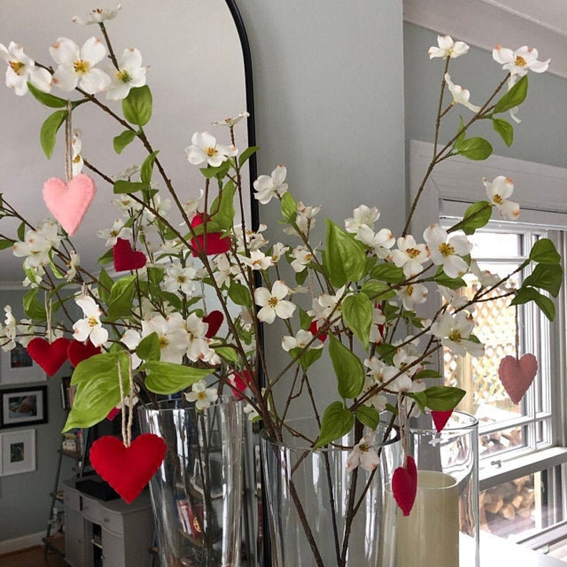 May include: A bouquet of white dogwood branches with felt hearts hanging from the branches. The branches are in two clear glass vases.